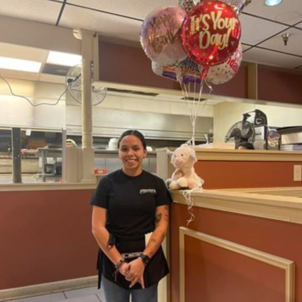 A smiling Jordana's Pizzeria employee stands beside a stuffed animal and birthday balloons.