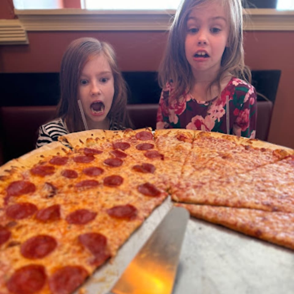 Two young girls with wide eyes and open mouths looking at a large pepperoni and cheese pizza.