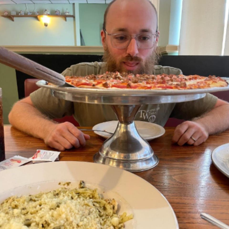 A man with a beard and glasses looks at a meat-topped pizza on a stand, with pasta in the foreground.