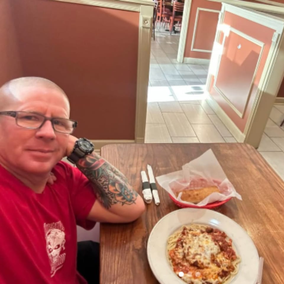 A man with a tattooed arm and glasses sits at a table with spaghetti and garlic bread.