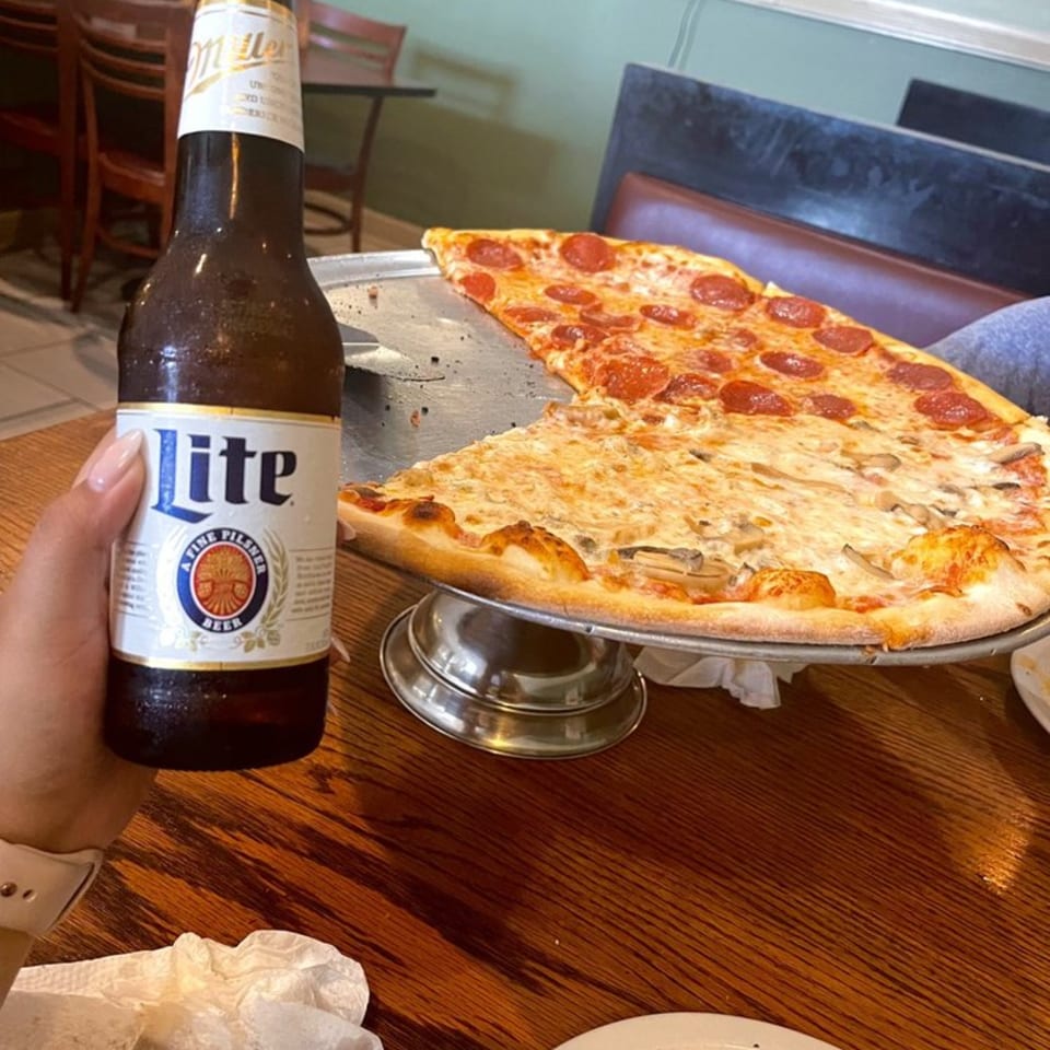 Hand holding a Miller Lite beer bottle next to a half-pepperoni, half-mushroom pizza on a stand.