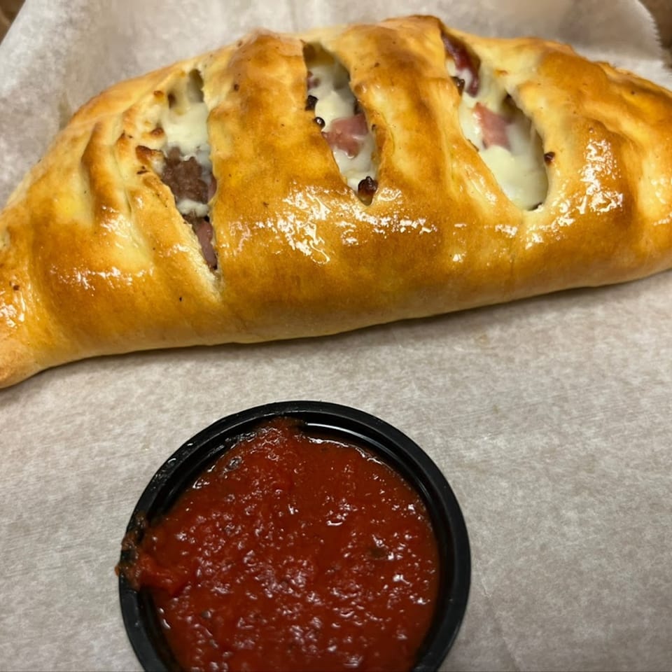 Golden-brown calzone with visible cheese and meat filling, served with a side of red marinara sauce.